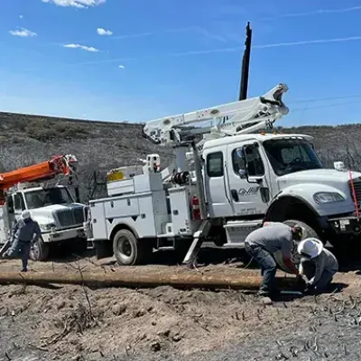Utility workers install a new wooden pole next to two service trucks in a barren, recently burned landscape under a clear sky.